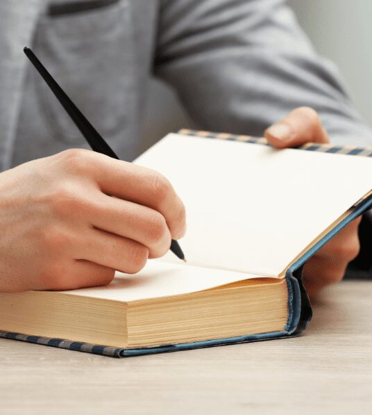 A person writing notes in a thick book with a pen.