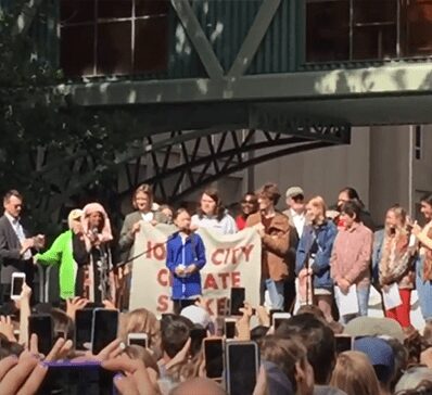 A crowd gathers around a speaker holding a 'No Climate Silence' sign.